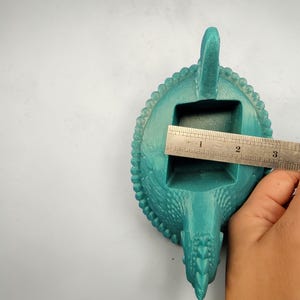 Puede incluir: Un cuenco para baratijas con forma de drag&oacute;n de color turquesa. El cuenco tiene una abertura rectangular, un borde con cuentas y una cabeza y cola de drag&oacute;n. La abertura mide aproximadamente 5 cm de ancho. El cuenco est&aacute; hecho de un material texturizado.