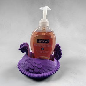 Puede incluir: Un soporte para dispensador de jab&oacute;n con forma de gallina morada con una botella transparente de jab&oacute;n de manos Softsoap Antibacteriano. El jab&oacute;n es de color naranja. El soporte est&aacute; hecho de un material texturizado y tiene un dise&ntilde;o detallado.