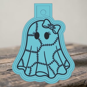 May include: A light blue cookie cutter shaped like a ghost with a stitched patchwork pattern, two oval eyes with eyelashes, and a bow on the head. The ghost figure features a small heart detail in the center of the bow and a top tab with a hole.