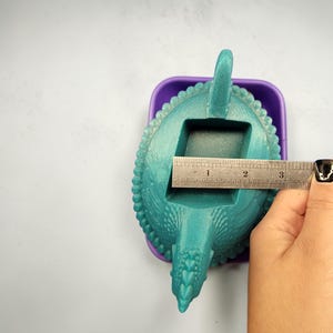 Puede incluir: Un molde en forma de dinosaurio de color turquesa con una superficie texturizada, dentro de un recipiente morado. El molde mide aproximadamente 7,6 cm de largo. Tiene una abertura rectangular, una cola y una cabeza.