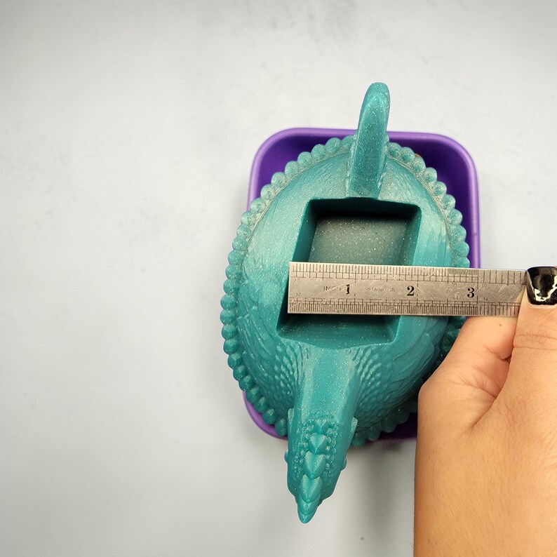 Puede incluir: Un molde en forma de dinosaurio de color turquesa con una superficie texturizada, dentro de un recipiente morado. El molde mide aproximadamente 7,6 cm de largo. Tiene una abertura rectangular, una cola y una cabeza.