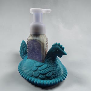Puede incluir: Un soporte para dispensador de jab&oacute;n con forma de gallina de color verde azulado. El soporte est&aacute; hecho de un material texturizado y presenta un dise&ntilde;o detallado de una gallina. Una botella con bomba blanca de jab&oacute;n de manos espumoso se coloca dentro.