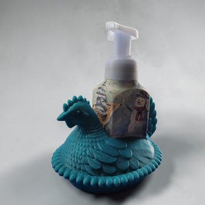 Puede incluir: Un dispensador de jab&oacute;n con forma de gallina de color turquesa. El dispensador tiene una escena invernal con un mu&ntilde;eco de nieve y un ciervo. La bomba es blanca.