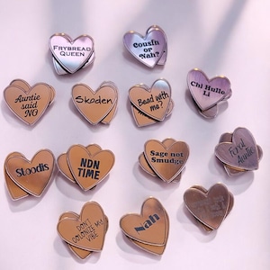 May include: A collection of heart-shaped pins in various colors, including silver, gold, and bronze. Each pin features a unique phrase or word, such as "Frybread Queen," "Cousin or Nah?" and "Don't Colonize My Vibe." The pins are arranged on a white surface.