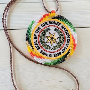 May include: A beaded pendant necklace featuring the Seal of the Cherokee Nation. The circular pendant has a white background with text and a star design, surrounded by bands of color. The necklace has a brown leather cord.