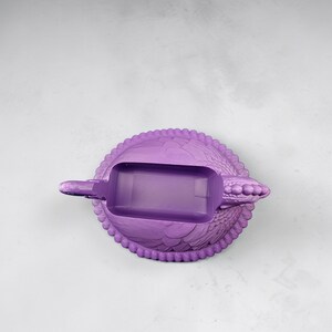 Puede incluir: Un cenicero morado con forma de tortuga. Tiene una abertura rectangular para las cenizas. El caparaz&oacute;n de la tortuga tiene un patr&oacute;n texturizado y el borde est&aacute; adornado con peque&ntilde;as cuentas redondas. Un objeto decorativo.