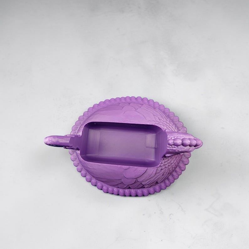 Puede incluir: Un cenicero morado con forma de tortuga. Tiene una abertura rectangular para las cenizas. El caparaz&oacute;n de la tortuga tiene un patr&oacute;n texturizado y el borde est&aacute; adornado con peque&ntilde;as cuentas redondas. Un objeto decorativo.