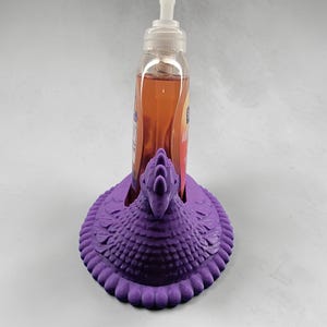 Puede incluir: Un soporte decorativo morado para dispensador de jab&oacute;n con forma de p&aacute;jaro. El soporte est&aacute; hecho de un material texturizado y presenta un dise&ntilde;o detallado. Una botella transparente de jab&oacute;n se coloca dentro del soporte.