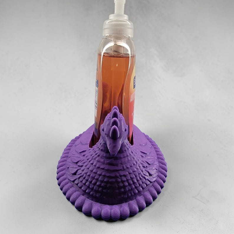 Puede incluir: Un soporte decorativo morado para dispensador de jab&oacute;n con forma de p&aacute;jaro. El soporte est&aacute; hecho de un material texturizado y presenta un dise&ntilde;o detallado. Una botella transparente de jab&oacute;n se coloca dentro del soporte.