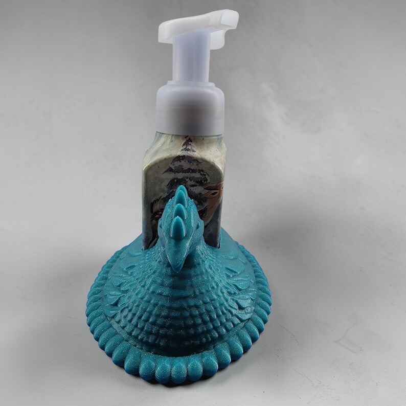 Puede incluir: Un dispensador de jab&oacute;n de color turquesa con una base en forma de pavo. El dispensador tiene una bomba blanca y una etiqueta con una escena invernal. La base del pavo tiene detalles de plumas y cabeza.