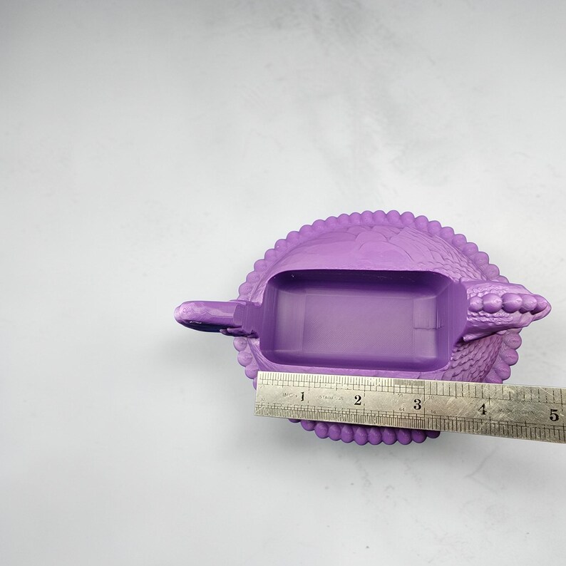 Puede incluir: Un cenicero morado con un dise&ntilde;o texturizado similar a un reptil. El cenicero tiene un compartimento rectangular y un peque&ntilde;o reposo extendido. Una regla muestra que el cenicero mide aproximadamente 10 cm de largo.