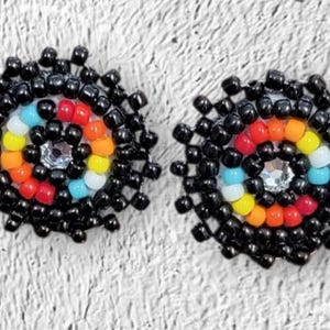 May include: A pair of handcrafted beaded stud earrings. The earrings feature a circular design with a central clear gem surrounded by a spiral of colorful beads in red, orange, yellow, white, and blue, all framed by black beads.