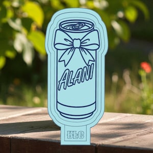May include: A light blue, can-shaped decorative item with a bow design and the name "ALANI" in a script font. The bottom of the item has the letters "BEC". The item is outdoors on a wooden surface.