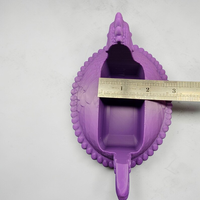 Puede incluir: Un recipiente morado con forma de drag&oacute;n con un interior rectangular y un asa. El recipiente mide aproximadamente 7,6 cm de largo. El drag&oacute;n tiene una superficie texturizada detallada y un borde decorativo.