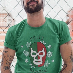 May include: A green t-shirt featuring a white wrestling mask graphic and the text "Feliz Navidad" in black. The mask is green, red, and white with a skull design. The shirt is surrounded by white snowflakes.