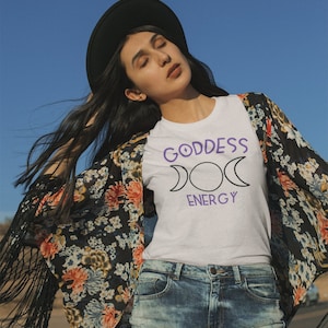 Może przedstawiać: Biała koszulka z fioletowym napisem "GODDESS ENERGY" oraz wzorem półksiężyca. Osoba ma na sobie dżinsowe szorty, czarny kapelusz i kwiatowe kimono z frędzlami.