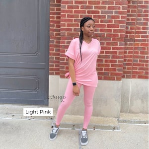 Op de afbeelding: Een lichtroze twee-delige outfit met een lang mouwloos topje en leggings. Het topje heeft een V-hals en een strik in de taille. De leggings zijn strak en hebben een rechte pijp. De outfit is gemaakt van een zachte, comfortabele stof. "Covered Couture" staat in de linkerbenedenhoek van de afbeelding. "Light Pink" staat in de linkerbenedenhoek van de afbeelding.