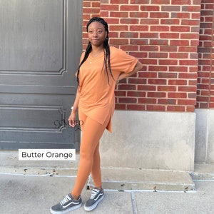 Op de afbeelding: Een tweedelige outfit in een botergele kleur. De outfit bestaat uit een shirt met korte mouwen en een V-hals en een legging. Het model draagt de outfit en staat voor een bakstenen muur.