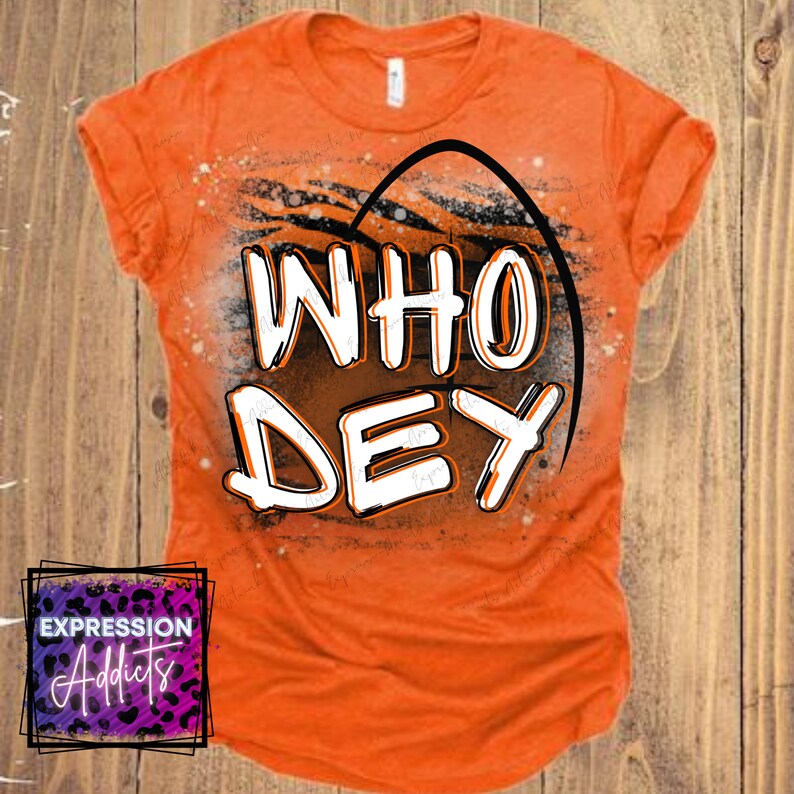 Puede incluir: Camiseta naranja con el texto "WHO DEY" en letras blancas y naranjas, delineadas en negro. El dise&ntilde;o incluye un arco negro y un patr&oacute;n de rayas de tigre. La camiseta tambi&eacute;n presenta el logotipo "Expression Addicts".