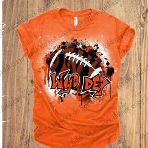 May include: Orange t-shirt featuring a football with tiger stripes and the words "Wild Day" in a graffiti style. The design includes black and white splatter effects, creating a bold, sporty look.