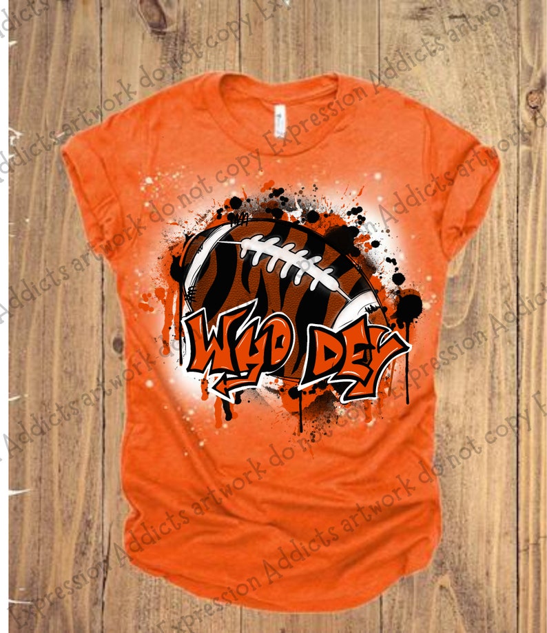 May include: Orange t-shirt featuring a football with tiger stripes and the words "Wild Day" in a graffiti style. The design includes black and white splatter effects, creating a bold, sporty look.