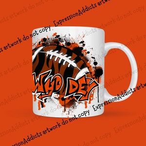May include: White ceramic mug featuring a football design with orange and black stripes, and the words "Wild Day" in graffiti style. The mug has a white handle and is decorated with orange paint splatters.