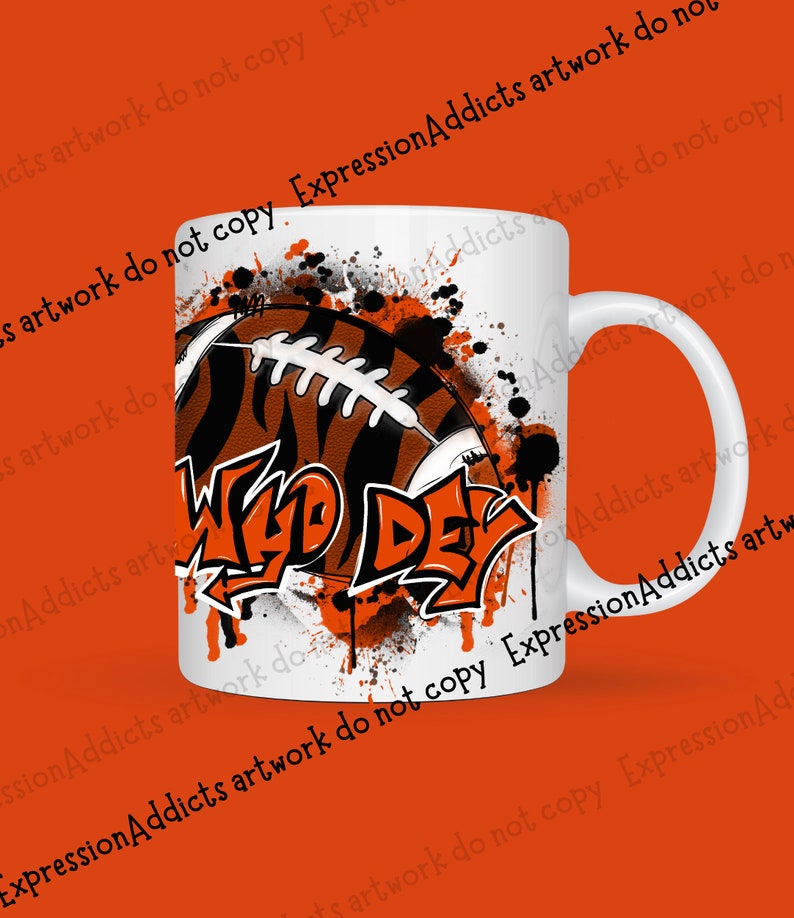 May include: White ceramic mug featuring a football design with orange and black stripes, and the words "Wild Day" in graffiti style. The mug has a white handle and is decorated with orange paint splatters.