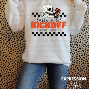 May include: White sweatshirt with the text "classy until KICKOFF" in orange and black, featuring a skull with a coffee cup and Cincinnati Bengals branding. The sweatshirt is paired with blue jeans.