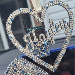 Könnte beinhalten: Ein herzförmiger Auto-Anhänger mit dem Namen "Hailey", der mit Strasssteinen ausgeschmückt ist. Der Anhänger ist mit klaren und blauen Strasssteinen verziert und hat einen kleinen, klaren, kristallartigen Anhänger.