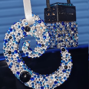 Puede incluir: Un ambientador de coche azul y plateado con pedrería y una perla negra colgando de una cinta blanca. El ambientador tiene forma de espiral y tiene el texto "Car Perfume" en la parte superior.