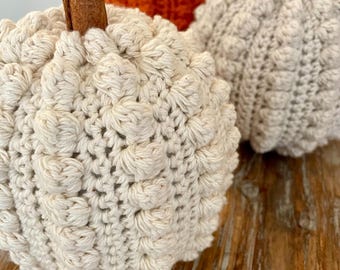 May include: Three handmade crochet pumpkins featuring a textured bobble stitch pattern. Two pumpkins are off-white and one is bright orange. Each pumpkin is topped with a brown cinnamon stick acting as a stem, creating a rounded, soft appearance.