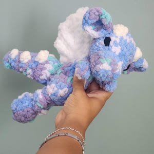 May include: A handmade crochet dragon toy in shades of blue, purple, and white. The plush dragon has a white wing and small black eyes. The toy is held in a person's hand against a light teal background.