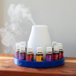 May include: A white essential oil diffuser with vapor rising, surrounded by small essential oil bottles in a blue holder. The bottles have white caps and colorful labels. The setup is on a wooden surface.