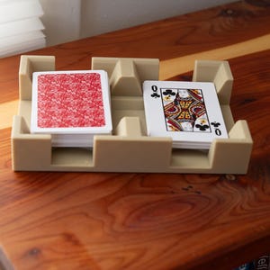 May include: A beige plastic playing card holder with two compartments, each holding a deck of cards. One deck has a red patterned back, and the other shows the Queen of Clubs. The holder sits on a wooden surface.