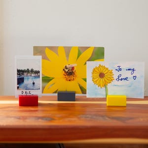 May include: A collection of framed art and photos displayed on a wooden surface. The images include a photo with the text "P.O.C.", a large yellow flower with a bee, and a watercolor painting of a sunflower with the text "To my Love".