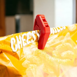 May include: A red plastic bag clip is attached to a yellow bag of cheesy snacks. The bag has the word "Cheesy" printed on it.