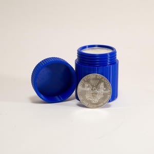May include: A blue, cylindrical container with a matching lid, next to a silver coin. The container is open, revealing a white substance inside. The coin displays an eagle and text, indicating it is from the United States.