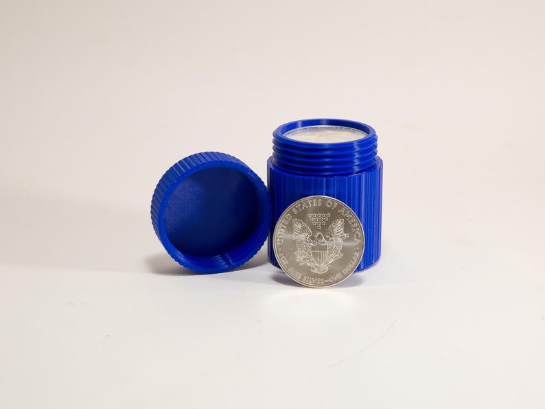 May include: A blue, cylindrical container with a matching lid, next to a silver coin. The container is open, revealing a white substance inside. The coin displays an eagle and text, indicating it is from the United States.
