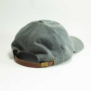 May include: A grey cotton baseball cap with a brown leather strap and a brass buckle. The strap has a small metal logo on it.