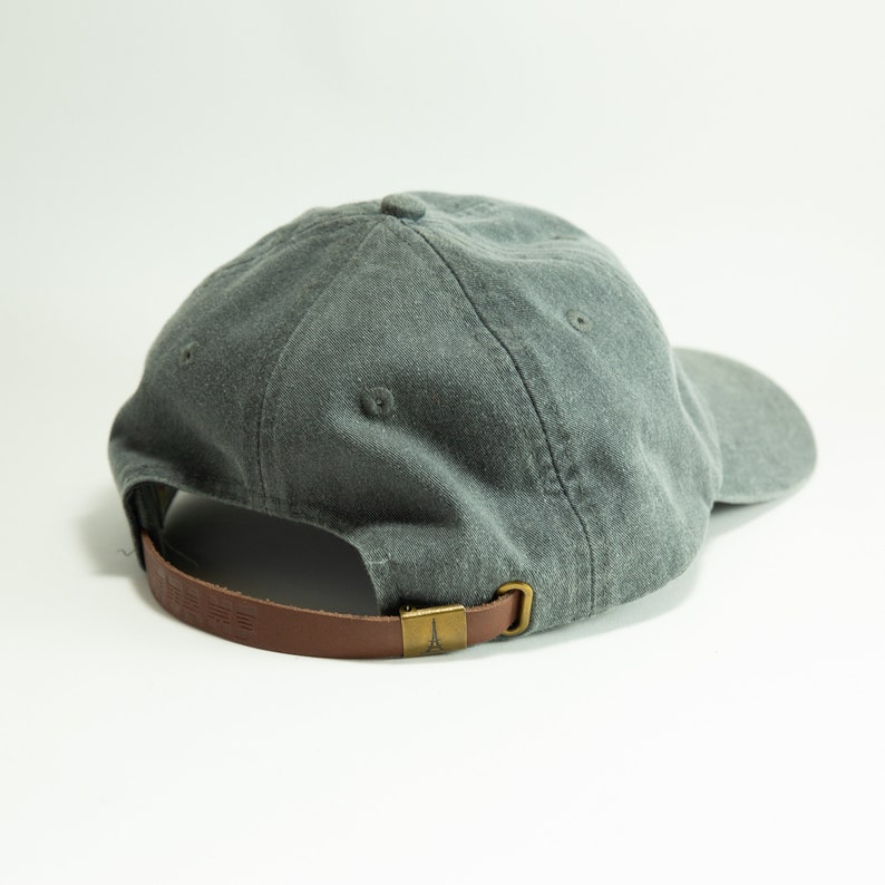 May include: A grey cotton baseball cap with a brown leather strap and a brass buckle. The strap has a small metal logo on it.