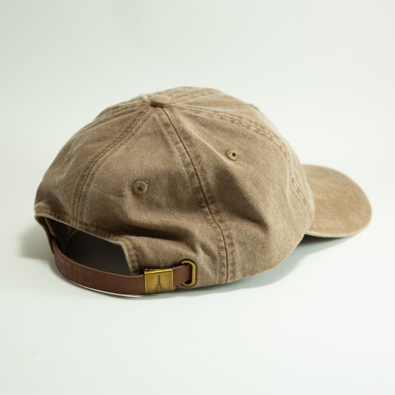 May include: A brown canvas baseball cap with a brown leather strap and a brass buckle. The buckle features a small Eiffel Tower design.