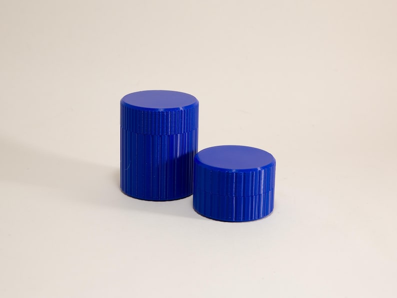 May include: Two cobalt blue cylindrical containers with ribbed sides and flat tops. One container is taller than the other. The containers are set against a plain, off-white background.