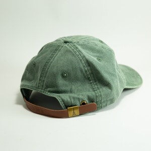 May include: A green cotton baseball cap with a brown leather strap and a gold metal buckle. The strap has a small Eiffel Tower design on it.