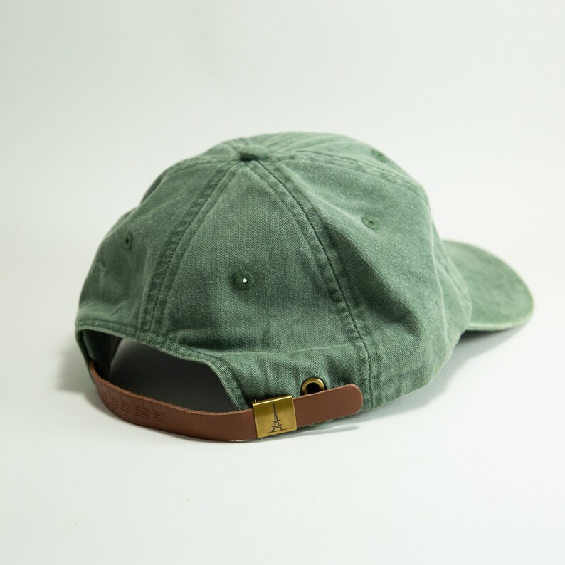 May include: A green cotton baseball cap with a brown leather strap and a gold metal buckle. The strap has a small Eiffel Tower design on it.