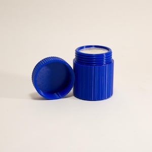 May include: A cobalt blue container with a matching lid. The cylindrical container has vertical ridges and a silver-colored interior. The lid is off to the side, revealing the contents. The items are on a white surface.