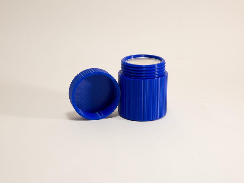May include: A cobalt blue container with a matching lid. The cylindrical container has vertical ridges and a silver-colored interior. The lid is off to the side, revealing the contents. The items are on a white surface.