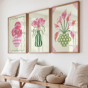 May include: Three framed floral art prints. Each print features a different arrangement of pink flowers in a decorative vase. The frames are a light brown colour, and the background is a neutral cream colour. The prints are displayed above a wooden bench with white cushions.