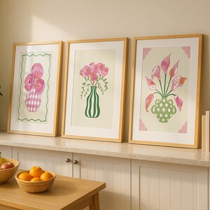 May include: Three framed floral art prints with wooden frames. Each print features a different arrangement of pink flowers in a vase. The prints are displayed on a white cabinet with a wooden table in front, holding fruit.