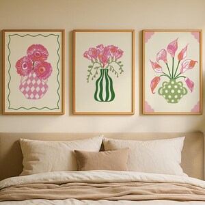 May include: Three framed floral art prints. The prints feature pink flowers in decorative vases. The vases have unique designs, including a diamond pattern, vertical stripes, and polka dots. The frames are a light wood colour, and the art is displayed above a bed.