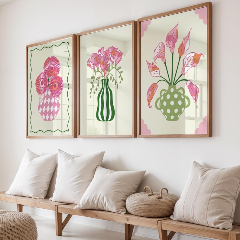 May include: Three framed floral art prints. Each print features a different arrangement of pink flowers in a decorative vase. The frames are a light brown colour, and the background is a neutral cream colour. The prints are displayed above a wooden bench with white cushions.
